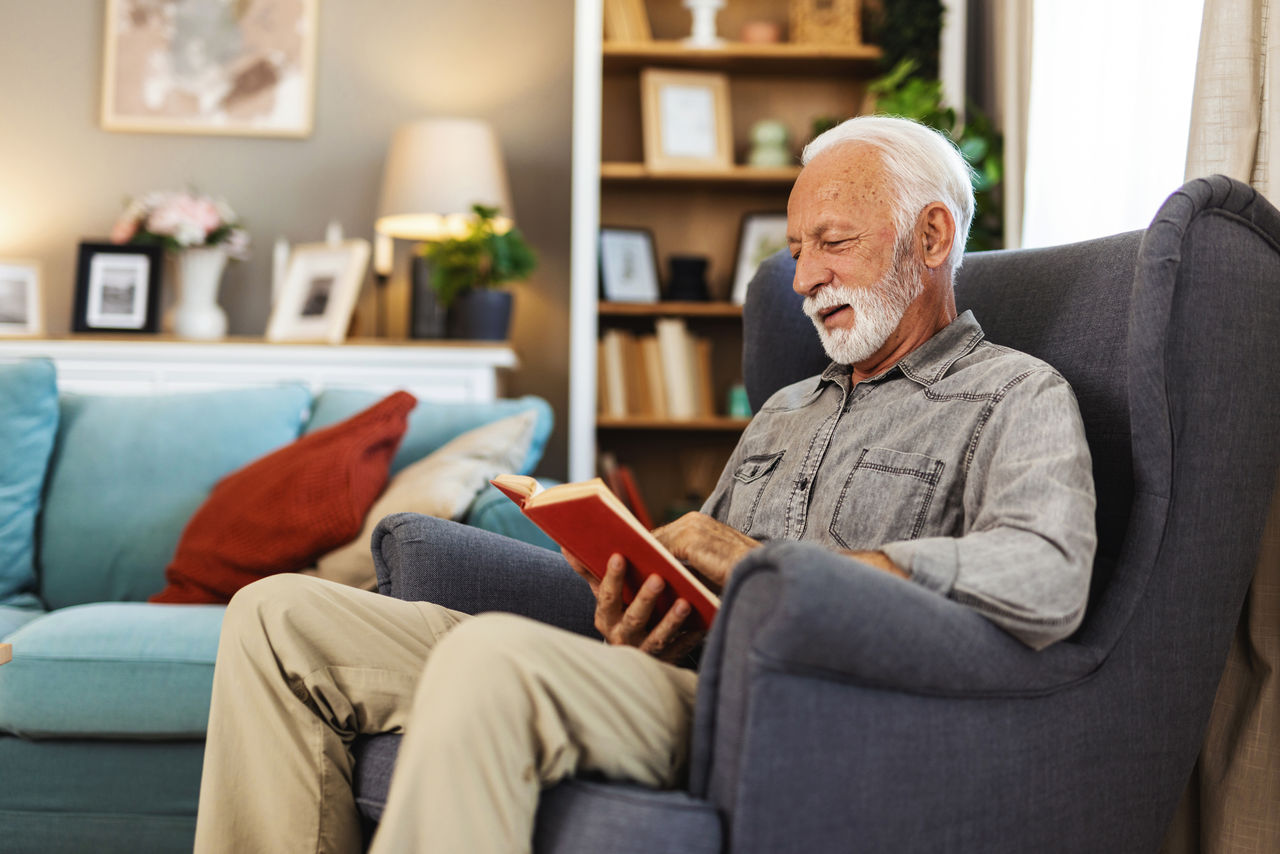 Senior man enjoys reading book alone in the living room. Old man alone at home.